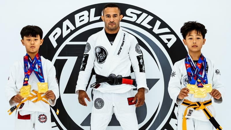 Two young grapplers wearing yellow belts beside a black belt in an IBJJF archive photograph.