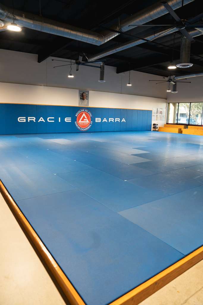 Wide interior photo of a Gracie Barra gym mat area with blue mats, wall padding, and windows.