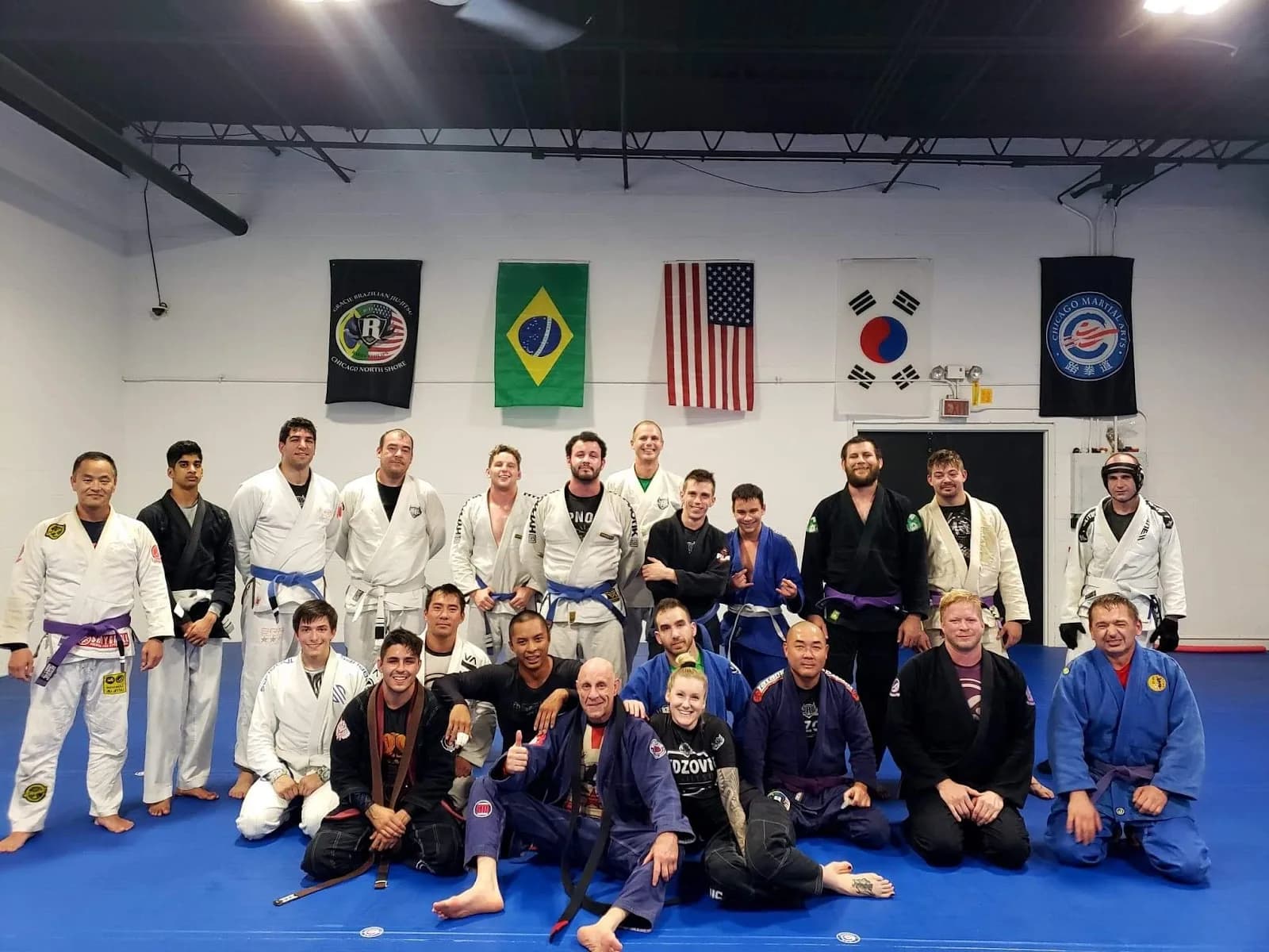 Group of adults in Brazilian Jiu-Jitsu gis and rashguards posed on blue mats inside a gym, flags (Brazil, USA, South Korea) and banners hanging on a white wall behind them.