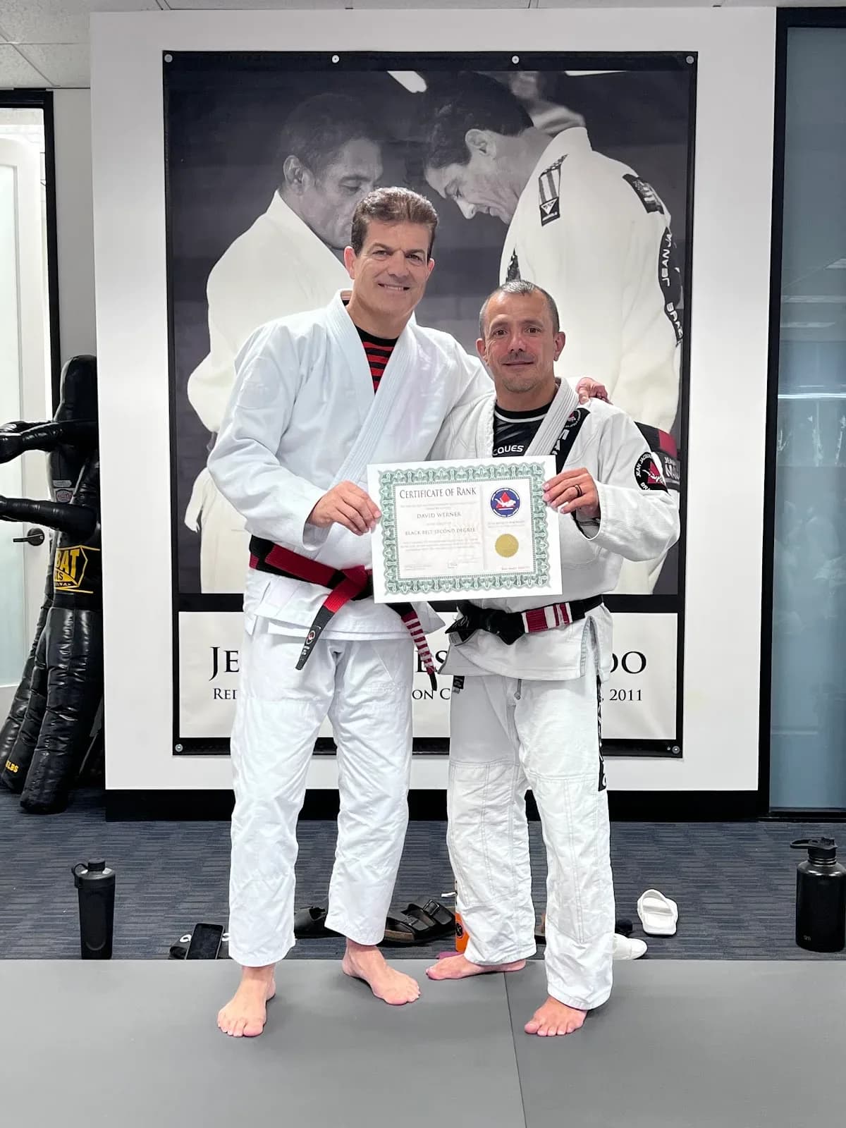 Two people in white gi standing on mats inside a gym, holding a framed certificate in front of a large black-and-white poster on the wall; gym gear and water bottles on the floor.
