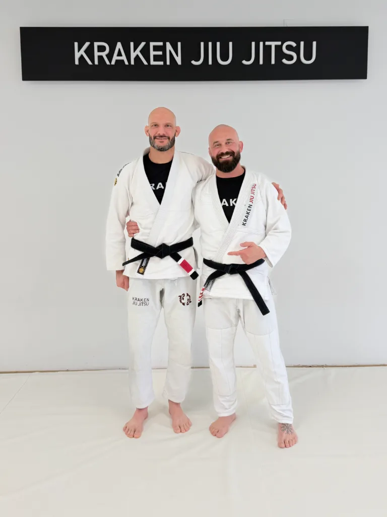 Two instructors in gis standing on mats under a gym sign reading 'KRAKEN JIU JITSU'.