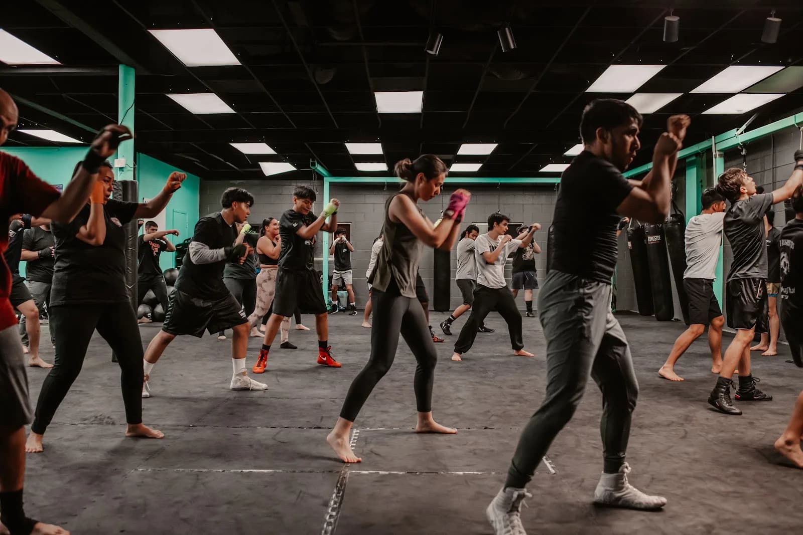 Large indoor martial arts class at Switch Fitness San Diego El Cajon: many people practicing striking footwork and punches on a matted floor with teal support columns and hanging heavy bags.