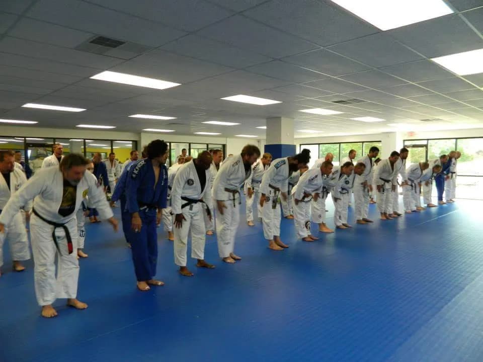 Large group of people in white and blue gis lined up on blue mats bowing toward the front of a bright martial arts training room at R&D Academy of Self Defense, Winston-Salem, NC.