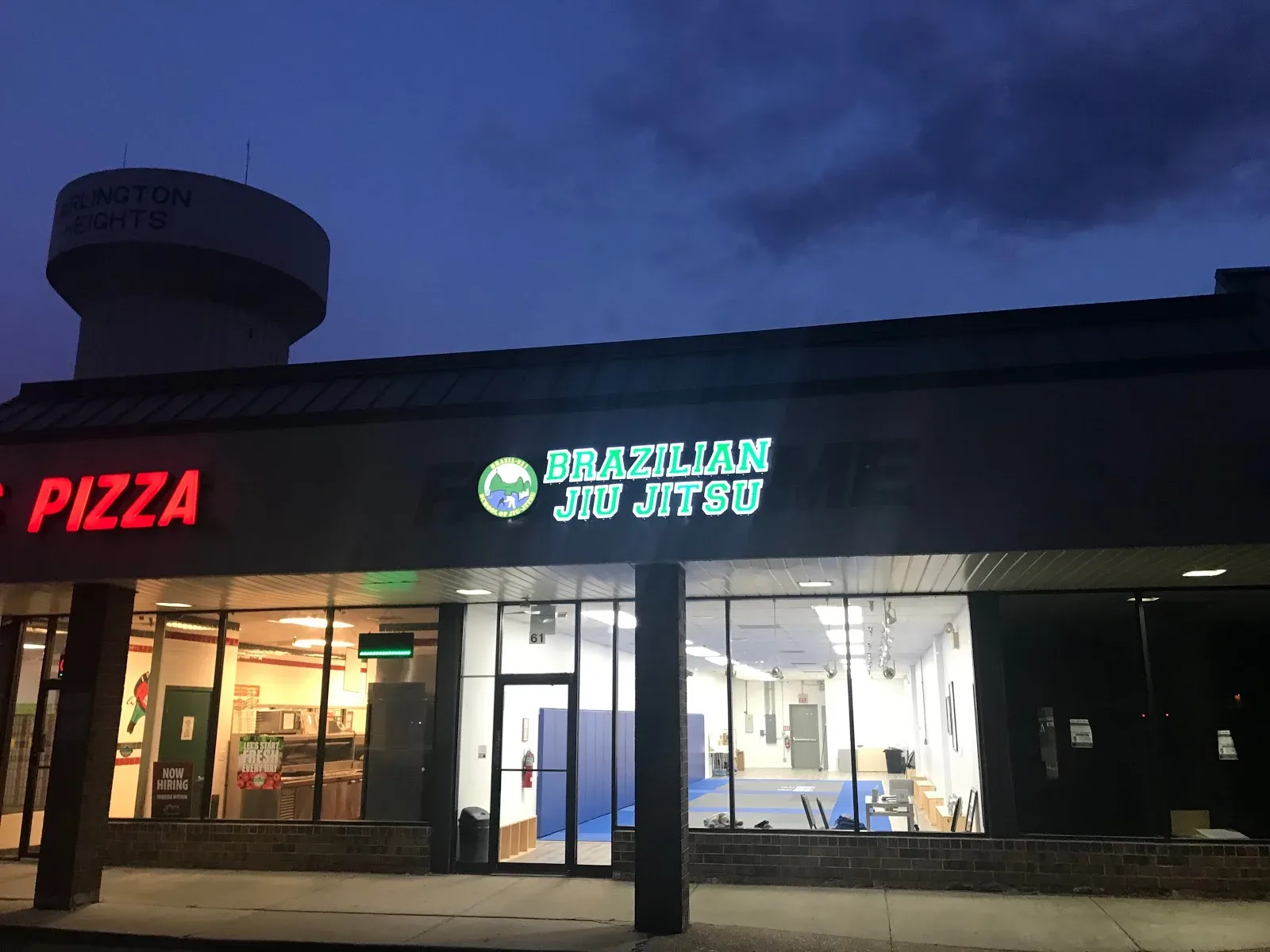 Street-front nighttime photo of Brazil 021 Arlington Heights storefront with illuminated green sign reading 'BRAZILIAN JIU JITSU' above large windows showing blue mats inside; neighboring pizza sign at left and a water t