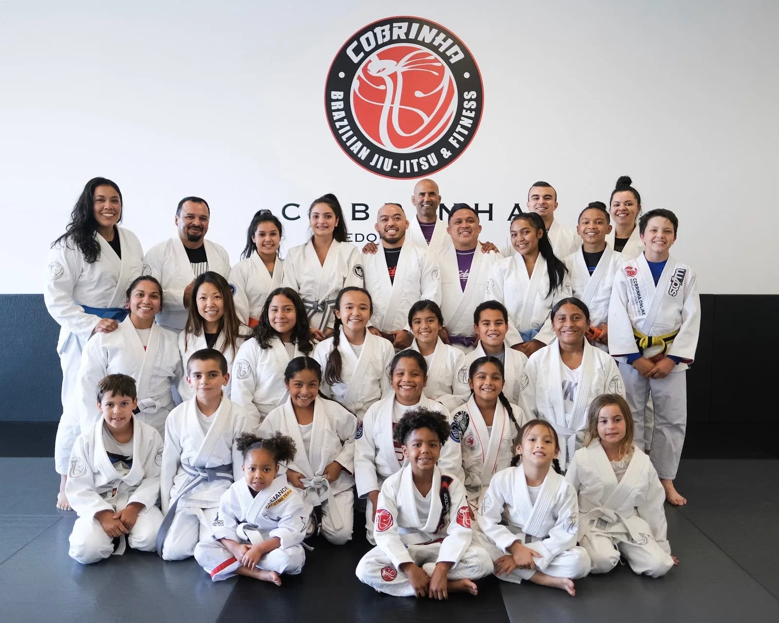 Group of adults and children in white jiu-jitsu gis posed in rows on mats under a large circular Cobrinha Brazilian Jiu-Jitsu & Fitness logo on a white wall at Alliance Jiu-Jitsu South Bay (Lawndale, CA).
