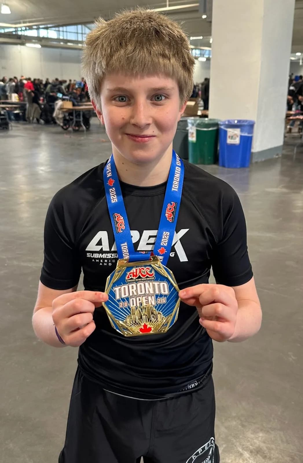 Person wearing an Apex Submission Grappling black rash guard and holding a large Toronto Open 2025 medal on a blue lanyard in an indoor event space.