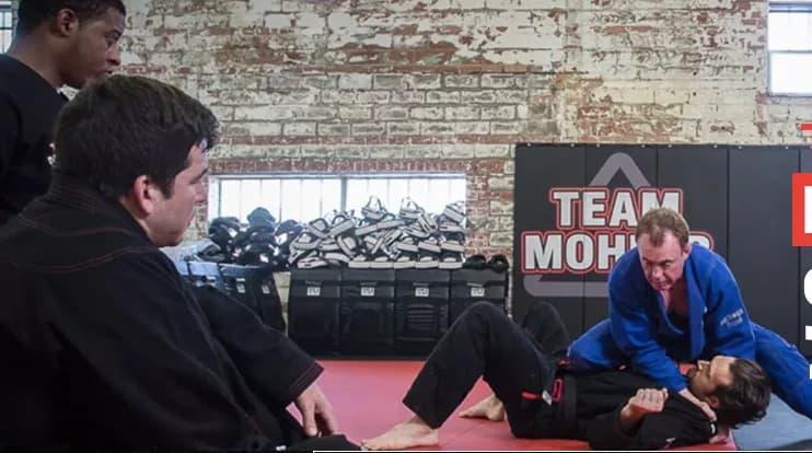 Training scene inside Mohler MMA gym showing instructors and students on red mats practicing Brazilian Jiu-Jitsu near a brick wall with 'TEAM MOHLER' signage.