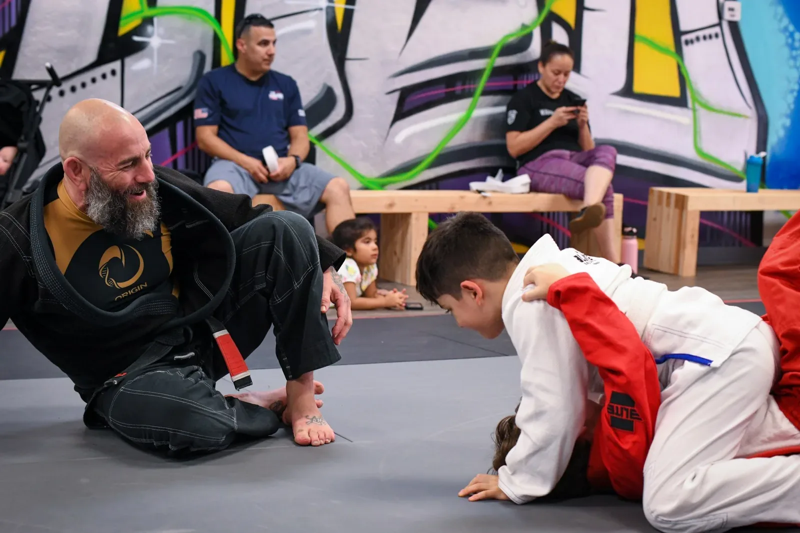Indoor gym scene at Undisputed Green Valley: adults and children on mats practicing Brazilian Jiu-Jitsu near colorful graffiti wall, with seated observers on wooden benches.