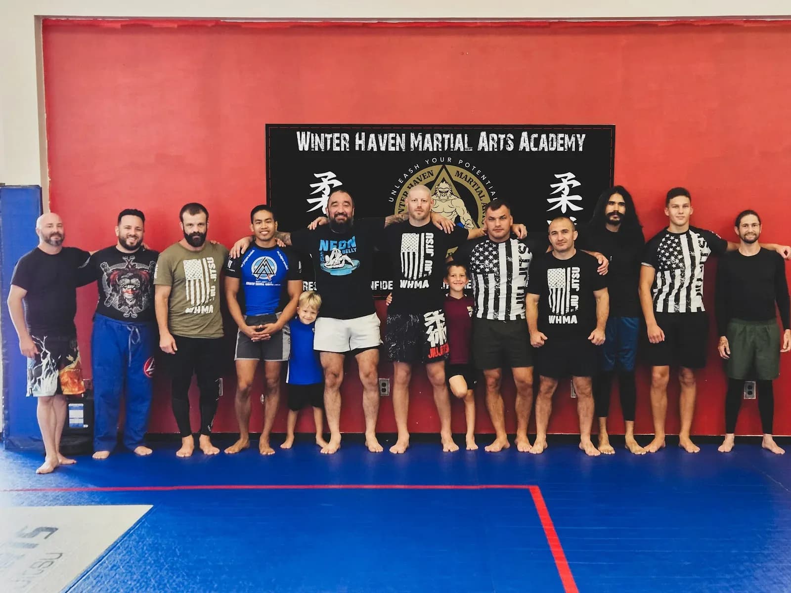 Group of adults and two children standing barefoot on blue mats in front of a red wall with a large black Winter Haven Martial Arts Academy banner.