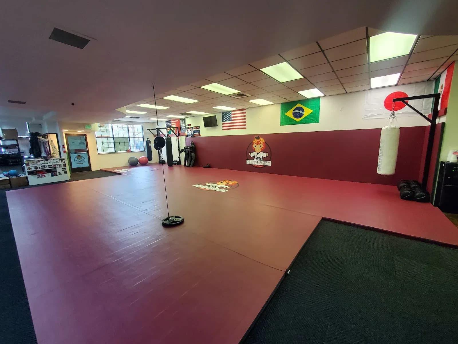 Interior photo of an empty martial arts training room with red mats, wall logo, hanging heavy bag, flags on the wall, and exercise equipment near the far wall.