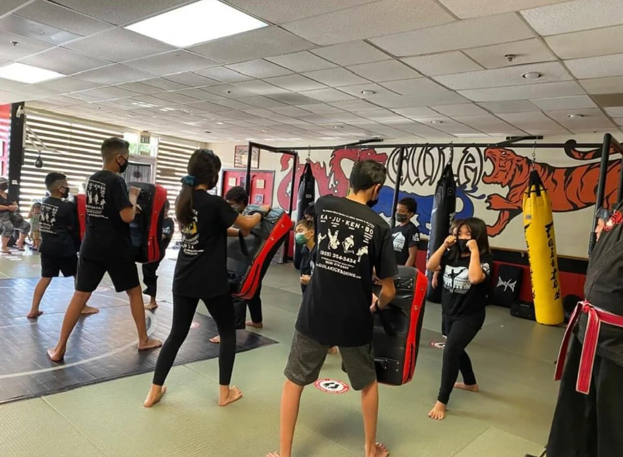 Children and teens in a martial arts gym practicing with rectangular kick shields and wearing black training shirts and face masks, padded mats and hanging heavy bags visible.