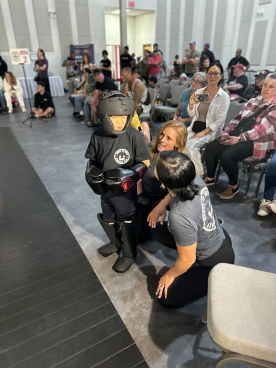 Coach kneeling beside a youth fighter in full sparring gear at a competition with seated spectators behind them