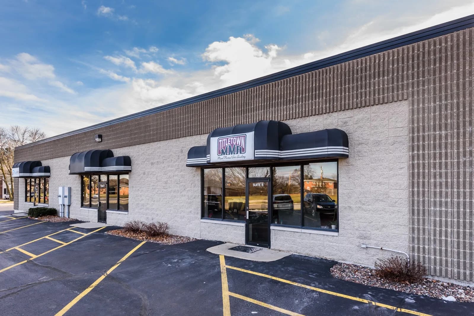Exterior storefront of Titletown MMA Academy in a single-story commercial building with black awnings and a sign above the entrance, parking spaces in front, Green Bay, WI.