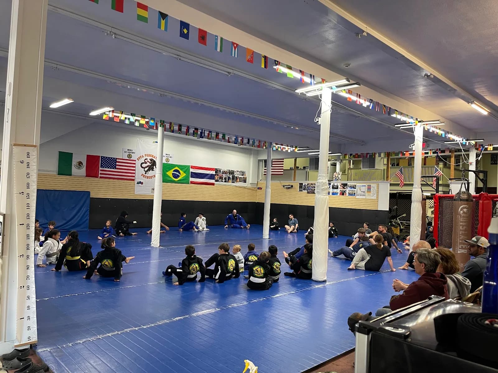 Interior view of Ferreira Family Fight Academy in Ukiah: blue matted training area with children and adults sitting in a circle, flags on the wall, punching bag and seating area.