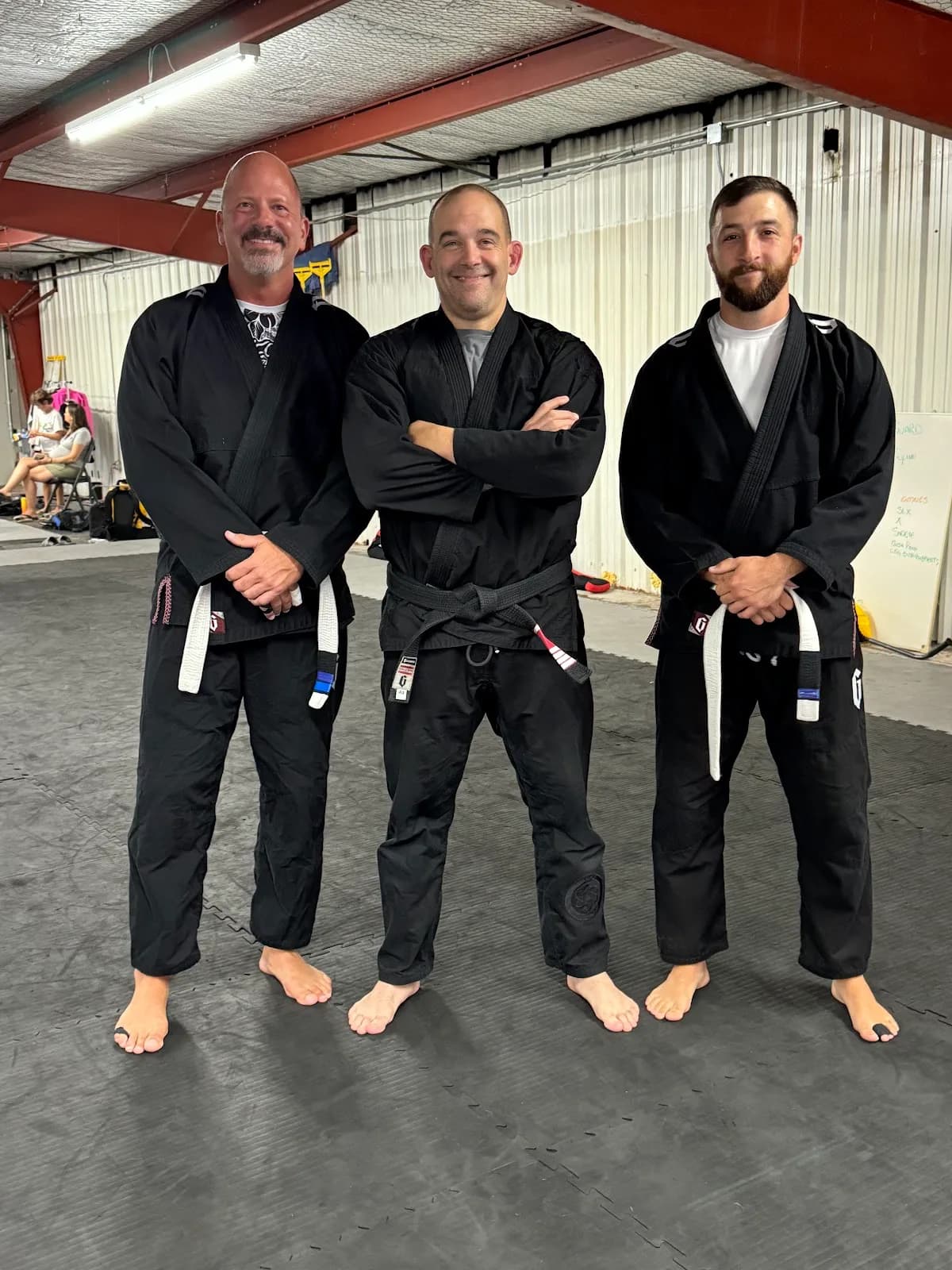 Three adults standing barefoot on black mats inside a metal-walled gym, wearing black gi uniforms with white belts visible; gym equipment and a whiteboard in the background.