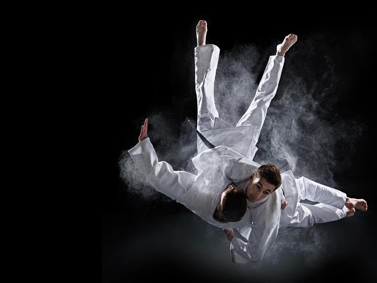 Two athletes in white gis performing a judo/jiu-jitsu throw on a dark background, powder or chalk visible in the air.
